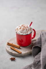 Red mug of hot cocoa or chocolate with whipped cream and cinnamon sticks on a gray background.