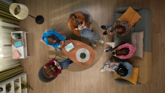 Top view of a group of female students in a living room sitting around a table on a couch and on chairs. Each of the girls is using her smartphone, looking at photos, videos, social media, schedules.