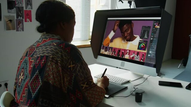 Black female photographer using graphic tablet and stylus pen while editing photos on computer during post-production process in studio