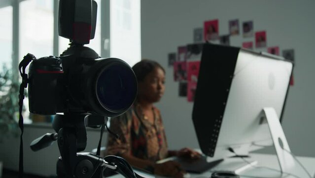 Selective focus shot of professional digital camera on tripod in photo studio, female photographer doing post-production in background