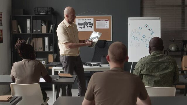 Rear view of unrecognizable diverse students listening to professor explaining shooting tactics using workbook during class in military college