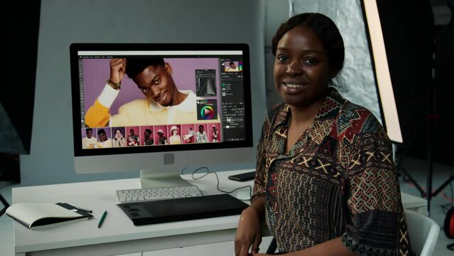 Portrait of cheerful African American female photographer sitting at desk with computer, posing for camera and smiling while editing images in studio