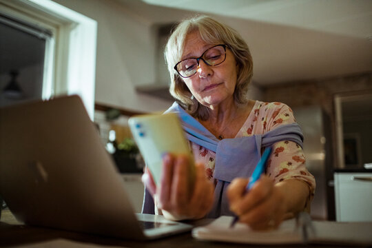 Senior woman taking notes on notebook from smartphone at home