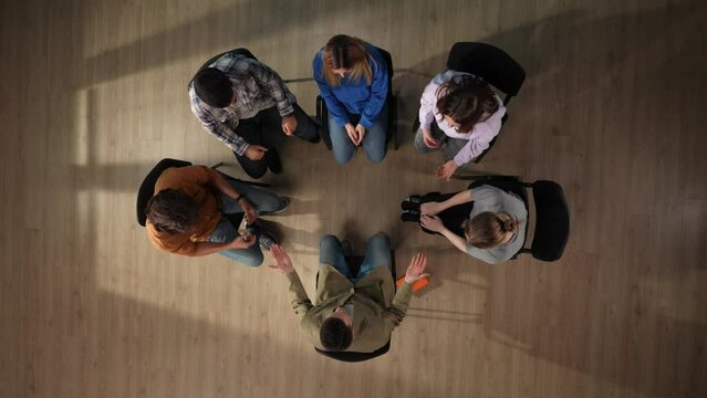 In the shot above, a group of people are sitting in a circle. They are having a therapy session with a psychologist and are sharing their problems. Then they hold hands raising them up and smiling