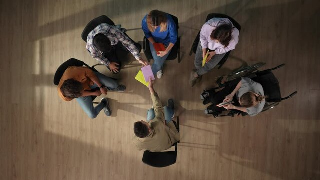 In the shot above, a group of people and their mentor are sitting on a chair. They have come for therapy. They are sitting in a circle writing something in a notebook with a disabled person