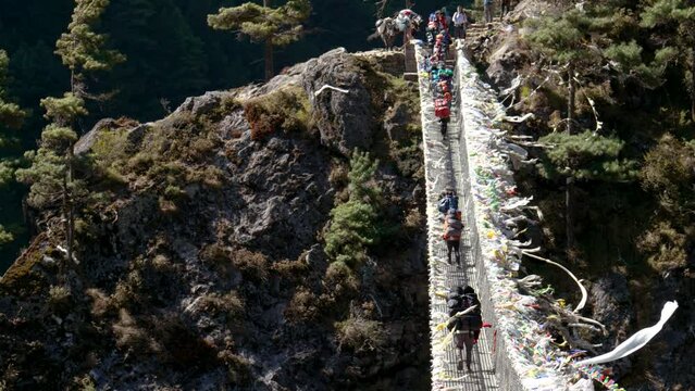 Trekking to Everest Base Camp. Tourists and sherpas with backpacks walk on Hillary suspension bridge through deep canyon of Himalayan mountains. Prayer flags and ribbons are fluttering in wind.