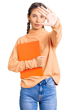 Beautiful caucasian woman with blonde hair holding book with open hand doing stop sign with serious and confident expression, defense gesture