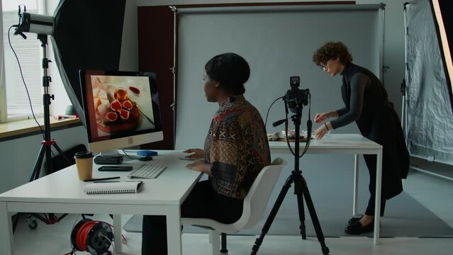 African American female photographer zooming image of figs on computer and giving instructions to food stylist working on sets in studio