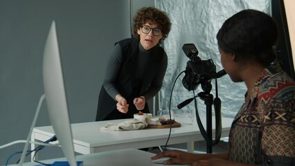 African American female photographer sitting at desk with computer in studio and talking to Caucasian stylist arranging food on table for camera before photoshoot