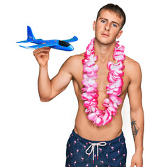Young blond man wearing swimsuit and hawaiian lei holding airplane toy thinking attitude and sober...