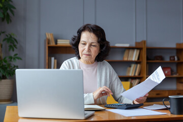 Middle aged senior woman sit with laptop and paper document. Pensive older mature lady reading...