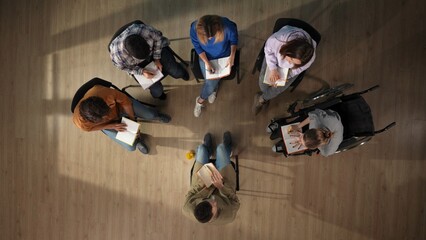In the shot above, a group of people and their mentor are sitting on a chair. They have come for therapy. They are sitting in a circle writing something in a notebook with a disabled person - obrazy, fototapety, plakaty