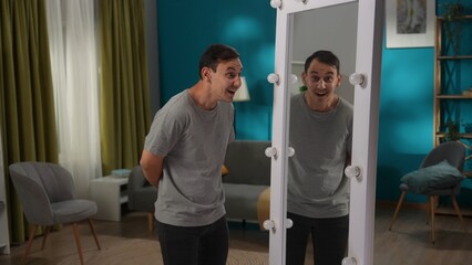 A man with bipolar disorder at a mirror in the living room. The man is writhing, laughing and mocking his reflection. Dark side, bipolar disorder, dissociative disorder.