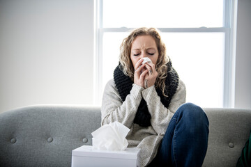 upset young woman sitting on sofa covered with blanket freezing blowing running nose got fever...