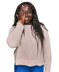 Young african woman wearing wool winter sweater covering one eye with hand, confident smile on face and surprise emotion.