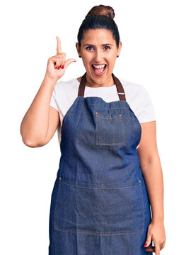 Young beautiful brunette woman wearing apron pointing finger up with successful idea. exited and happy. number one.