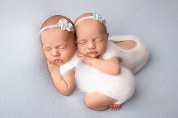 Tiny newborn twin girls. A newborn twin embraces and sleeps on his sister, Newborn twin girls against a pale blue blanket with white bandages and white delicate woolen bodysuits. 