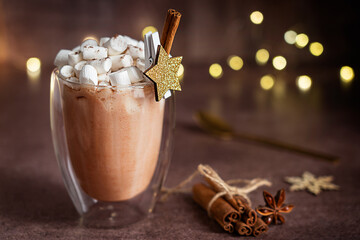 Homemade cocoa drink or hot chocolate with marshmallow topping decorated with cinnamon stick and wooden star decor served in glass cup or mug on brown table against christmas garland defocus lights