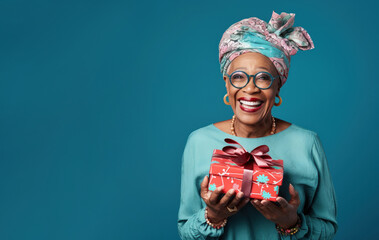 Beautiful African American mature woman 50, 60, 70 years old on a blue background holding a large gift box, holiday surprise, compliment bonus for birthday anniversary - obrazy, fototapety, plakaty