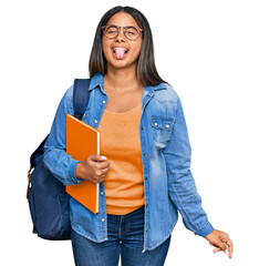 Young latin girl wearing student backpack and holding books sticking tongue out happy with funny expression. emotion concept.