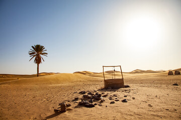 Old water well in the desert with palm tree and dunes in the background - obrazy, fototapety, plakaty