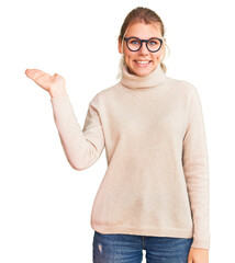 Young beautiful blonde woman wearing turtleneck sweater and glasses smiling cheerful presenting and pointing with palm of hand looking at the camera.