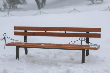 A snow-covered empty wooden bench stands in a park under white snow in a frosty winter. Photography, nature.