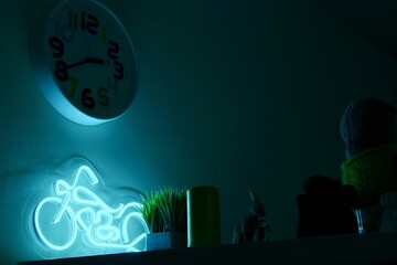 A neon night light in the shape of a motorcycle in the room illuminates the wall and table with a plant with a soft Blue light in the night. 