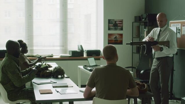 Side full shot of Caucasian military professor teaching how to use and hold shotgun and then explaining to diverse cadets individually during training class in service college