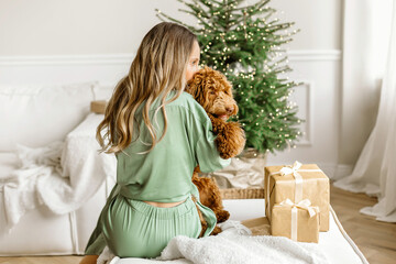 A Beautiful young woman kissing a golden brown labradoodle dog on christmas tree , bokeh light garland bacground. Cute Family puppy and girl play at home, new year decorated interior at Christmas  - obrazy, fototapety, plakaty