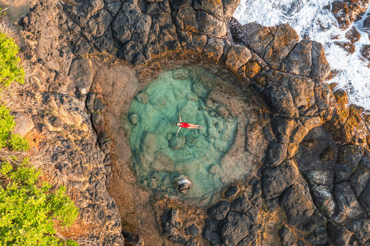 Aerial view of a woman swimming in a natural rock pool along the coastline with waves breaking on the cliffs near Gris Gris beach, Savanne, Mauritius.