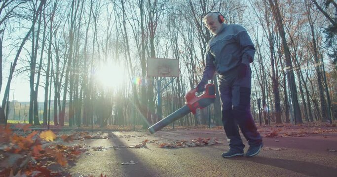 Aged caucasian man in overalls, headphones and gloves using electric blower for removing dry leaves at park. Janitor taking care of city area during autumn season.