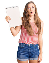 Beautiful caucasian young woman holding notebook scared and amazed with open mouth for surprise, disbelief face