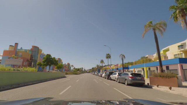 First person view, FPV, from dashcam of car driving along the Algarve Coast in Portugal in Albufeira, driving in the city by colourful buildings. Road trip video in POV, with blue sky