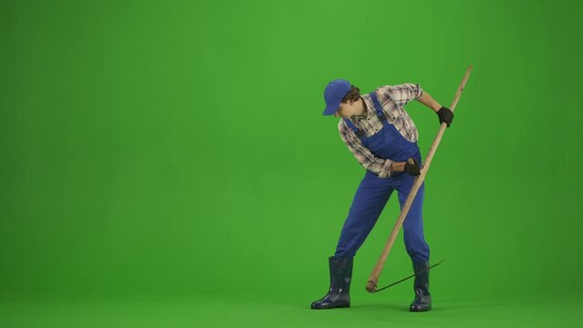 Portrait of male in overalls and rubber boots on chroma key green screen. Man gardener mowing the grass on the soil with an old scythe, looking positive.