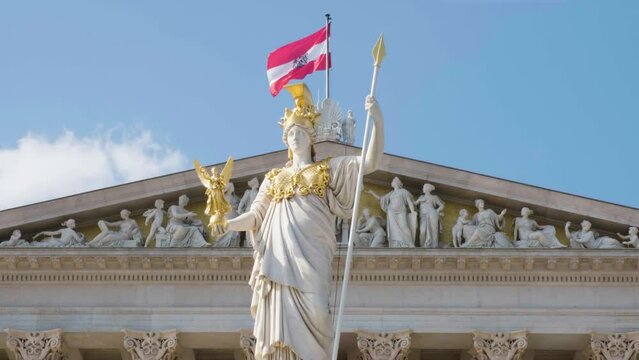 Austrian flag waving in the wind on top of the Wien Parliament building with statue in front.