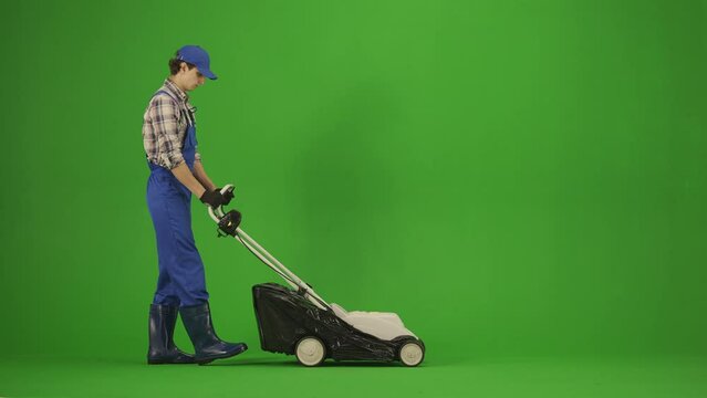 Portrait of male in overalls and rubber boots on chroma key green screen. Man gardener mowing his grass lawn with modern mower, tired expression.