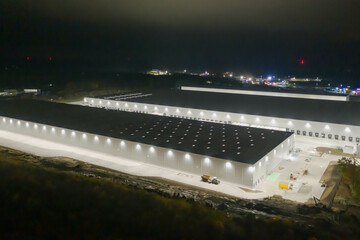 Centrum logistyczne w nocnych ciemnościach. Teren wokół jak i budynki oświetlone są sztucznym światłem elektrycznym. Zdjęcie zrobione przy użyciu drona. - 689822502