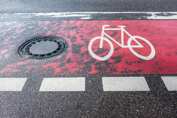 Radweg mit Symbol Fahrrad mit abgefahrenem rotem Fahrbahnbelag und Kanaldeckel