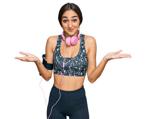 Beautiful brunette woman wearing gym clothes and using headphones clueless and confused expression...