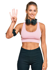 Young blonde woman wearing gym clothes and using headphones showing and pointing up with fingers number four while smiling confident and happy.
