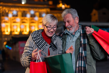 Cheerful senior couple at Christmas market buying gifts for family and friends looking into gift bag and smiling. - obrazy, fototapety, plakaty