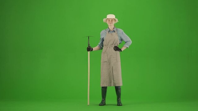 Portrait of female in apron and rubber boots on chroma key green screen. Woman gardener stands holding hoe in hand and looking at camera smiling.