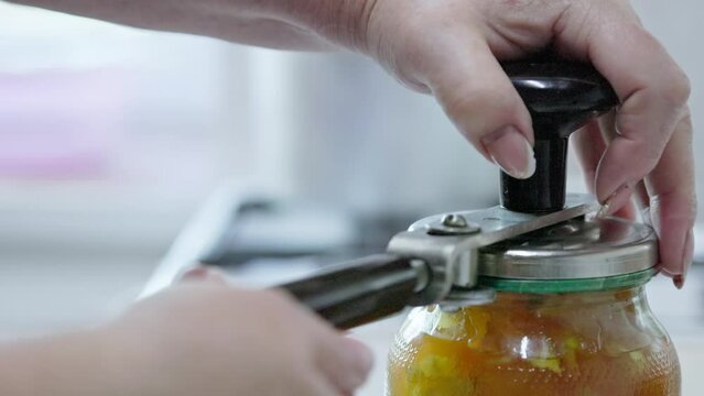 A woman cans apple jam at home. Closes the glass jar with a metal lid with rolling tool. Slow motion closeup.