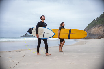 Cheerful surfer couple, walking along the shore with their surfboards.