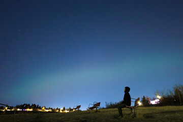 Man sitting on bench admiring the faint glow of the northern lights at Bowmont Park in Calgary