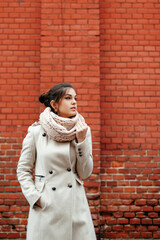 Beautiful brunette girl in white coat and scarf standing against brick wall and looking away