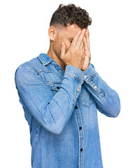 Young hispanic man wearing casual denim jacket with sad expression covering face with hands while...