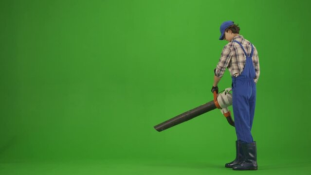 Portrait of male in overalls and rubber boots on chroma key green screen. Man gardener cleans the ground with garden vacuum leaf blower.