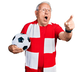 Senior man with grey hair football hooligan holding ball pointing with finger surprised ahead, open...